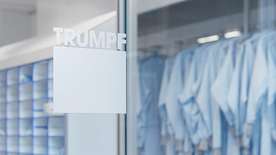Innenansicht eines industriellen Raums mit blauen Regalen und hängenden blauen Arbeitskleidungen. An der Glastür steht der Schriftzug “TRUMPF”.