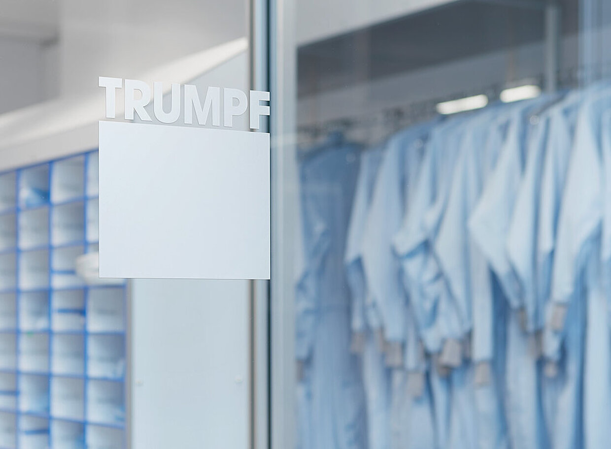 Industrieller Raum bei TRUMPF Innenansicht eines industriellen Raums mit blauen Regalen und hängenden blauen Arbeitskleidungen. An der Glastür steht der Schriftzug “TRUMPF”.