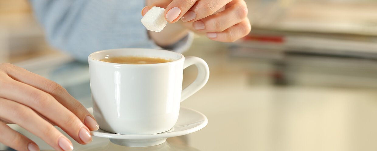 Nahaufnahme einer Hand, die einen Zuckerwürfel in eine Tasse Kaffee gibt, auf einem Tisch in einem gemütlichen Raum.
