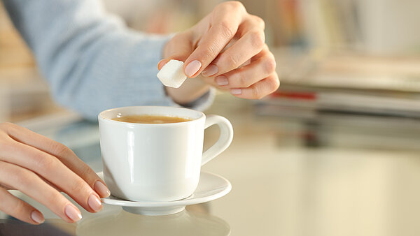 Nahaufnahme einer Hand, die einen Zuckerwürfel in eine Tasse Kaffee gibt, auf einem Tisch in einem gemütlichen Raum.