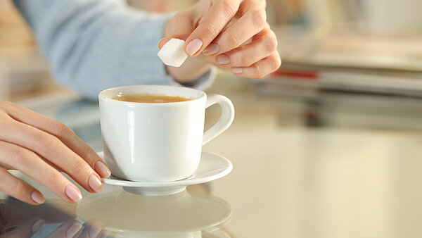 Nahaufnahme einer Hand, die einen Zuckerwürfel in eine Tasse Kaffee gibt, auf einem Tisch in einem gemütlichen Raum.