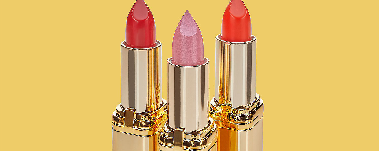 Drei Lippenstifte in Rot, Rosa und Orange mit goldener Hülle vor gelbem Hintergrund.