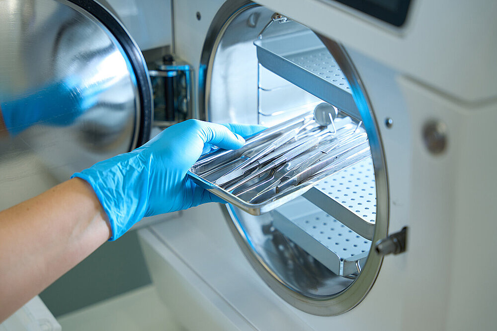 Eine Hand in OP-Handschuh schiebt ein Tray mit medizinischen Werkzeugen in einen Autoklav