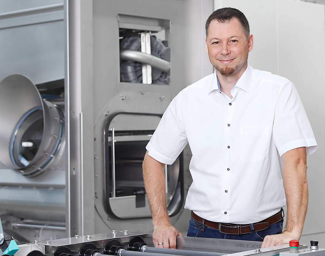 Mitarbeiter Stefan Schaal vor einer Maschine Ein Mann in einem weißen Hemd steht in einer Produktionsanlage neben einer Maschine.