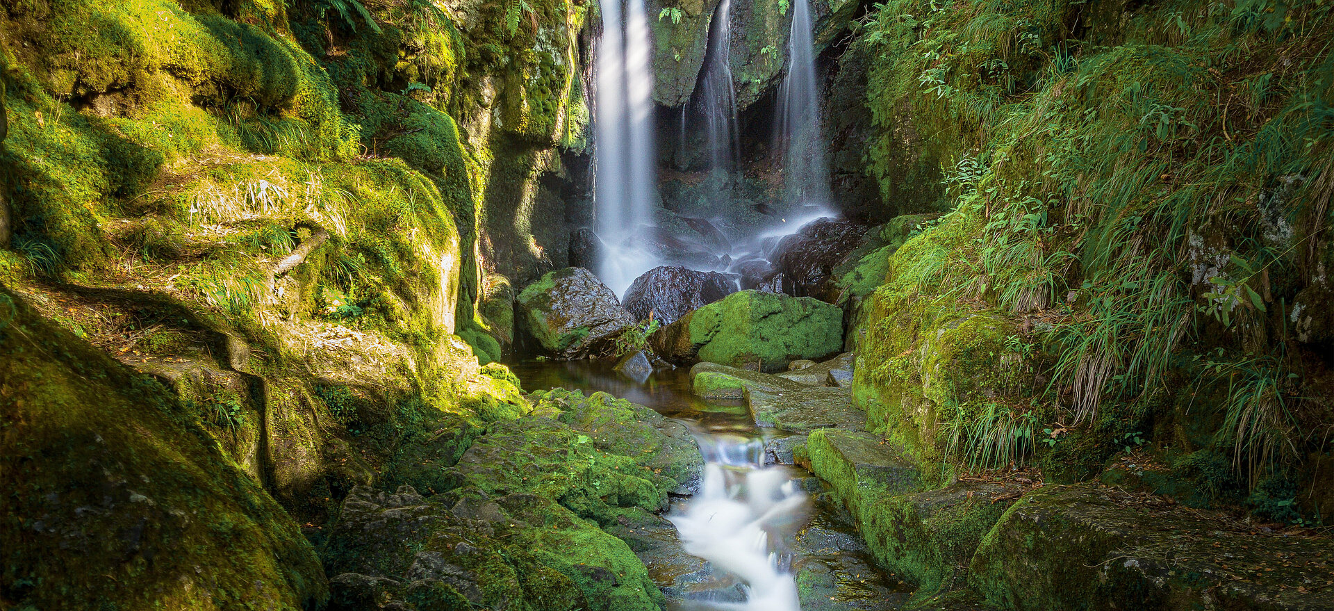 Ein idyllischer Wasserfall fließt über moosbewachsene Felsen in einem grünen Wald, symbolisierend die Kraft und Reinheit der Natur.