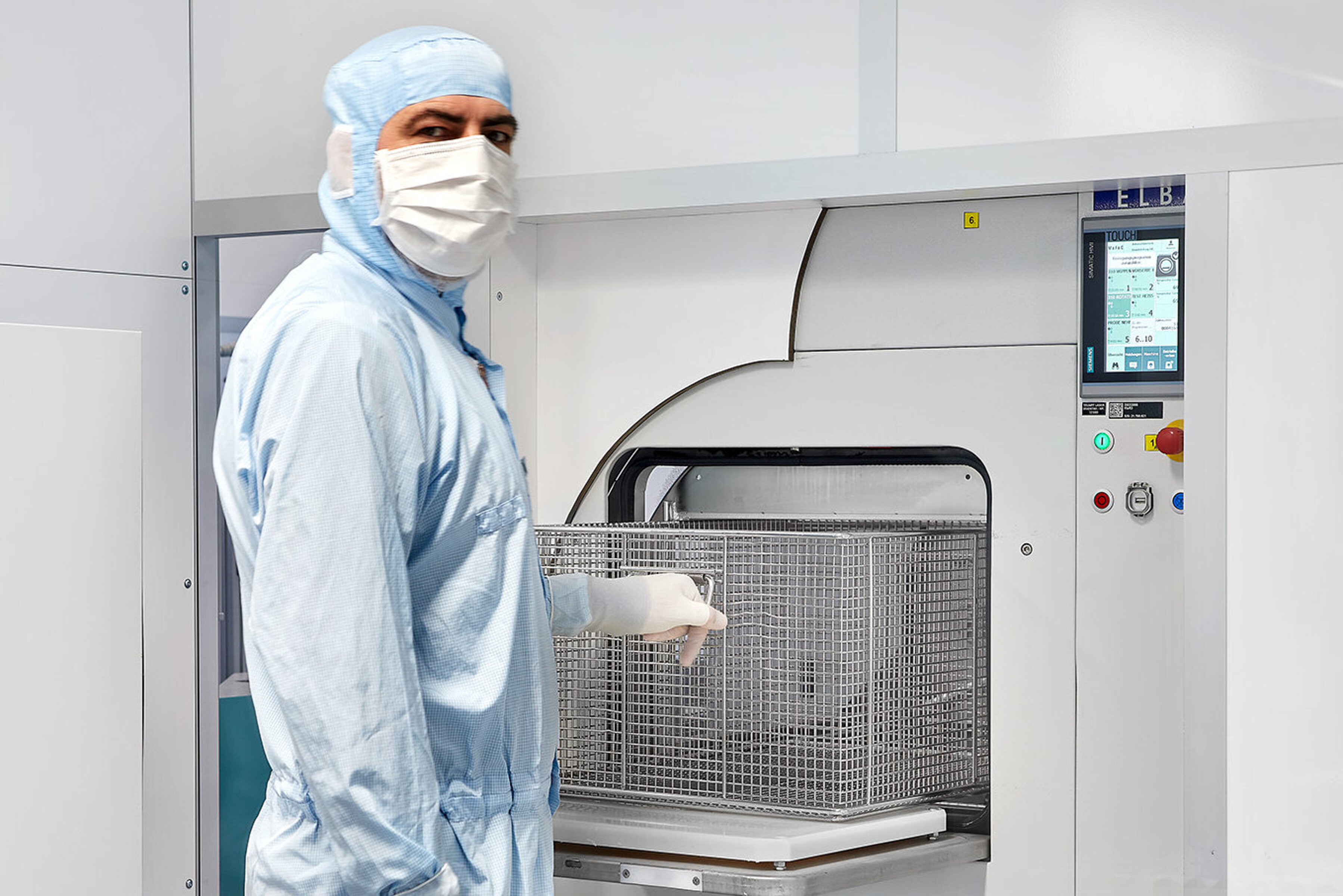 Bedienung einer industriellen Reinigungsanlage ELBA mit Schutzkleidung. Person in Schutzkleidung bedient eine industrielle Reinigungsanlage und lädt einen Metallkorb ein.
