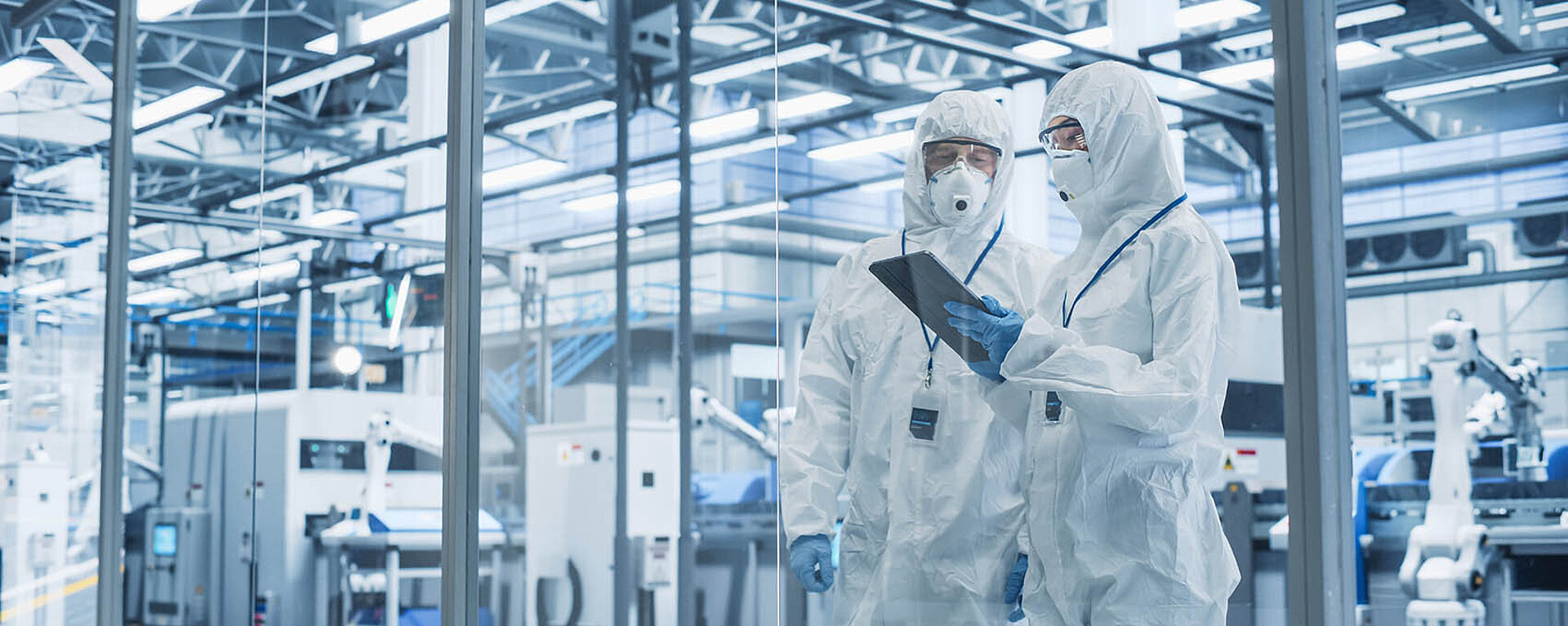 Zwei Personen in Schutzkleidung stehen in einer modernen industriellen Anlage und betrachten ein Tablet.